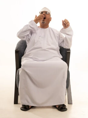 الشعور بالإرهاق والتعب، التثاؤب والحاجة إلى النوم، وضع اليد على الفم بإيماءات الشعور بالنعاس، بورتريه لرجل كبير مسن عربي عماني خليجي مبتسم يرتدي الدشداشة والكمه ونظارات طبية يجلس على كرسي مريح، خلفية بيضاء.