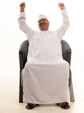 رفع اليدين إلى الأعلى بإيماءات الشعور بالنعاس، الشعور بالإرهاق والتعب، بورتريه لرجل كبير مسن عربي عماني خليجي مبتسم يرتدي الدشداشة والكمه ونظارات طبية يجلس على كرسي مريح، خلفية بيضاء.