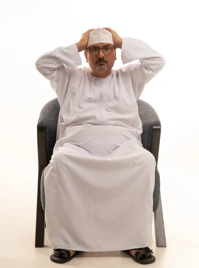 تلقي أخبار صادمة ومفاجئة، النظر إلى الكاميرا بإيماءات الصدمة والذهول، بورتريه لرجل كبير مسن عربي عماني خليجي مبتسم يرتدي الدشداشة والكمه ونظارات طبية يجلس على كرسي مريح ويضع يديه فوق رأسه، خلفية بيضاء.