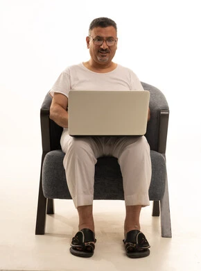 Accomplishing remote work tasks, a modern and advanced technical device, expressions of focus and contemplation, a portrait of an elderly Arab Saudi Gulf man wearing pants and a t-shirt and wearing reading glasses sitting on a comfortable chair using a laptop, white background.