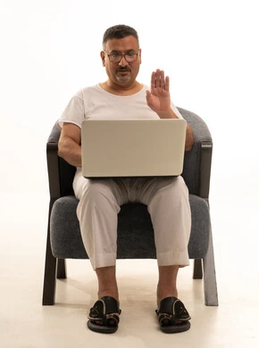 Raising the hand with gestures of greeting and peace, making a video call, completing remote work tasks, a modern and advanced technical device, a portrait of an elderly Arab Saudi Gulf man wearing pants and a t-shirt and wearing reading glasses sitting on a comfortable chair using a laptop, white background.