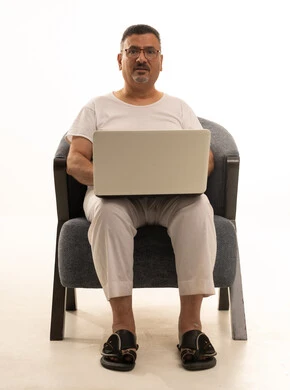 Accomplishing remote work tasks, a modern and advanced technical device, expressions of focus and contemplation, a portrait of an elderly Arab Saudi Gulf man wearing pants and a t-shirt and wearing reading glasses sitting on a comfortable chair using a laptop, white background.