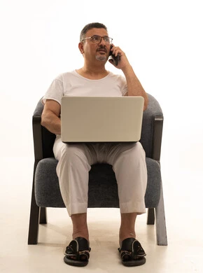 التواصل مع العملاء عبر الجوال، إجراء مكالمة هاتفية، إنجاز مهام العمل عن بعد، جهاز تقني حديث ومتطور، بورتريه لرجل كبير بالسن عربي سعودي خليجي يرتدي سروال و فانيلة ويضع نظارات طبية يجلس على كرسي مريح يقوم باستخدام الحاسوب المحمول، خلفية بيضاء