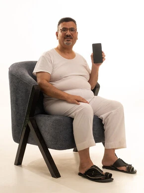 Browsing social media, displaying a blank black screen on a mobile phone, completing remote work tasks, a modern and advanced technical device, a portrait of an elderly Arab Saudi Gulf man wearing pants and a t-shirt and wearing reading glasses sitting on a comfortable chair using the phone, white background.