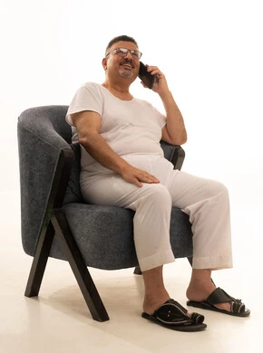Communicating with family and friends, enjoyment and entertainment using modern technology, making a phone call via mobile, a portrait of an elderly Arab Saudi Gulf man wearing pants and a t-shirt and wearing reading glasses sitting on a comfortable chair using a mobile phone, white background.