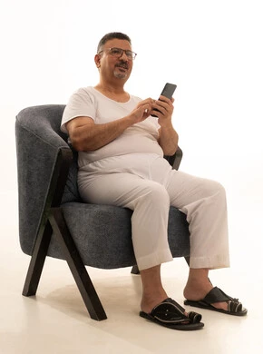 Enjoyment and entertainment using modern technology, browsing social media sites via mobile, completing work tasks remotely, a portrait of an elderly Arab Saudi Gulf man wearing pants and a t-shirt, wearing reading glasses, sitting on a comfortable chair using a mobile phone, white background.