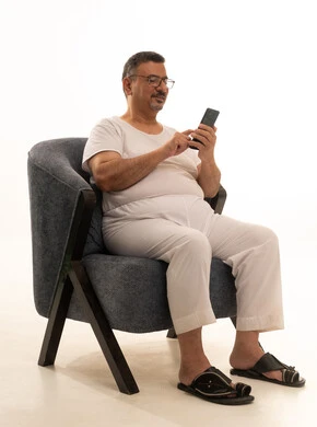 Enjoyment and entertainment using modern technology, browsing social media sites via mobile, completing work tasks remotely, a portrait of an elderly Arab Saudi Gulf man wearing pants and a t-shirt, wearing reading glasses, sitting on a comfortable chair using a mobile phone, white background.