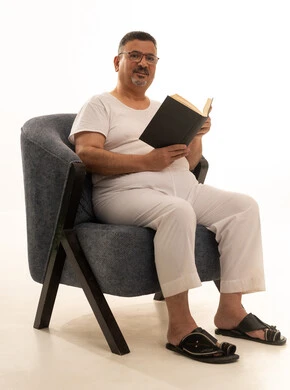 Reading enjoyable stories, the concept of self-learning, expressions of contemplation and focus, spending free time reading, learning, and culture, a portrait of an elderly Arab Saudi Gulf man wearing pants and a t-shirt sitting on a comfortable chair reading a book, acquiring information from books and novels, white background.
