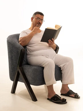 The concept of self-learning, expressions of contemplation and focus, spending free time reading, learning, and culture, a portrait of an elderly Arab Saudi Gulf man wearing pants and a t-shirt sitting on a comfortable chair reading a book, acquiring information from books and novels, white background.