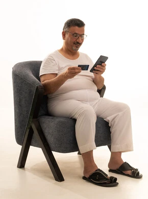 The possibility of paying bills with electronic cards, the ease of purchasing and paying with a bank credit card, a portrait of an elderly Arab Gulf Saudi man wearing pants and a shirt and medical glasses sitting on a comfortable chair holding an ATM card and working on a mobile phone, white background.