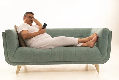 تصفح مواقع التواصل الاجتماعي عبر الجوال، جهاز تقني حديث ومتطور، بورتريه لرجل كبير بالسن عربي سعودي خليجي يرتدي سروال و فانيلة ويضع نظارات طبية يجلس على اريكة مريحة يقوم باستخدام الهاتف المحمول، خلفية بيضاء