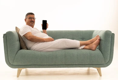 عرض شاشة سوداء فارغة عبر الجوال، جهاز تقني حديث ومتطور، بورتريه لرجل كبير بالسن عربي سعودي خليجي يرتدي سروال و فانيلة ويضع نظارات طبية يجلس على اريكة مريحة ينظر إلى الكاميرا ويحمل بيده الهاتف المحمول، خلفية بيضاء