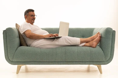 إنجاز مهام العمل عن بعد عبر اللابتوب، جهاز تقني حديث ومتطور، بورتريه لرجل كبير بالسن عربي سعودي خليجي يرتدي سروال و فانيلة ويضع نظارات طبية يجلس على اريكة مريحة يقوم باستخدام الحاسوب المحمول، خلفية بيضاء