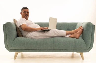 إنجاز مهام العمل عن بعد عبر اللابتوب، جهاز تقني حديث ومتطور، بورتريه لرجل كبير بالسن عربي سعودي خليجي يرتدي سروال و فانيلة ويضع نظارات طبية يجلس على اريكة مريحة يقوم باستخدام الحاسوب المحمول، خلفية بيضاء