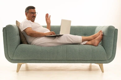 إجراء مكالمة فيديو، إنجاز مهام العمل عن بعد عبر اللابتوب، جهاز تقني حديث ومتطور، بورتريه لرجل كبير بالسن عربي سعودي خليجي يرتدي سروال و فانيلة ويضع نظارات طبية يجلس على اريكة مريحة يقوم باستخدام الحاسوب المحمول، خلفية بيضاء