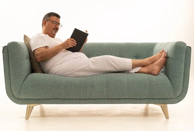 Gestures of contemplation and focus, spending free time reading, learning, and culture, the concept of self-directed learning, browsing books and novels, a portrait of an elderly Arab Saudi Gulf man wearing pants and a t-shirt sitting on a comfortable sofa reading a book, white background.