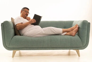 Gestures of contemplation and focus, spending free time reading, learning, and culture, the concept of self-directed learning, browsing books and novels, a portrait of an elderly Arab Saudi Gulf man wearing pants and a t-shirt sitting on a comfortable sofa reading a book, white background.
