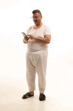 Browsing the internet via mobile, a modern and advanced technical device, accomplishing work tasks remotely, a portrait of an elderly Arab Saudi Gulf man wearing pants and a t-shirt and wearing glasses, using a mobile phone, full body, white background.