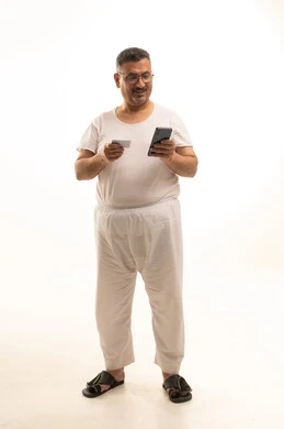 إمكانية تسديد الفواتير بالبطاقات الإلكترونية, استخدام جهاز تقني حديث, سهولة الشراء والدفع عن طريق بطاقة الإئتمان البنكية، بورتريه لرجل كبير السن عربي خليجي سعودي يرتدي سروال وفنيله يمسك بيده بطاقة الصراف ويعمل على الهاتف المحمول، الجسم بالطول الكامل, خلفية بيضاء