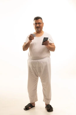 إمكانية تسديد الفواتير بالبطاقات الإلكترونية, استخدام جهاز تقني حديث, سهولة الشراء والدفع عن طريق بطاقة الإئتمان البنكية، بورتريه لرجل كبير السن عربي خليجي سعودي يرتدي سروال وفنيله يمسك بيده بطاقة الصراف ويعمل على الهاتف المحمول، الجسم بالطول الكامل, خلفية بيضاء