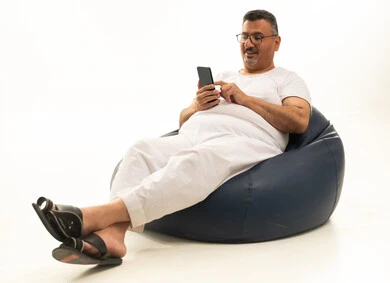 Browsing social media in free time, a modern and advanced technical device, a portrait of an elderly Arab Saudi Gulf man wearing pants and a t-shirt, wearing reading glasses, sitting on a chair between a comfortable bag, using a mobile phone, white background.