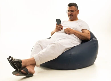 إجراء مكالمة فيديو، تصفح مواقع التواصل الاجتماعي، جهاز تقني حديث ومتطور، بورتريه لرجل كبير بالسن عربي سعودي خليجي يرتدي سروال و فانيلة ويضع نظارات طبية يجلس على كرسي بين باج مريح يقوم باستخدام الهاتف المحمول، خلفية بيضاء