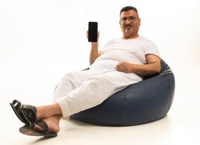 عرض شاشة سوداء فارغة عبر الجوال، جهاز تقني حديث ومتطور، بورتريه لرجل كبير بالسن عربي سعودي خليجي يرتدي سروال و فانيلة ويضع نظارات طبية يجلس على كرسي بين باج مريح ينظر إلى الكاميرا ويحمل بيده الهاتف المحمول، خلفية بيضاء