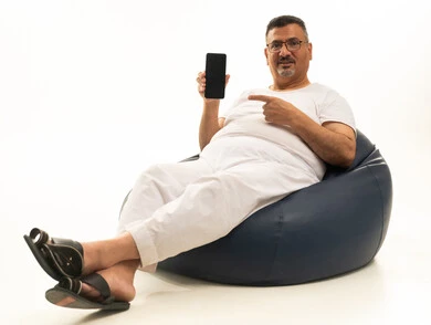 الاشارة بإصبع السبابة إلى شاشة سوداء فارغة عبر الجوال، جهاز تقني حديث ومتطور، بورتريه لرجل كبير بالسن عربي سعودي خليجي يرتدي سروال و فانيلة ويضع نظارات طبية يجلس على كرسي بين باج مريح ينظر إلى الكاميرا ويحمل بيده الهاتف المحمول، خلفية بيضاء