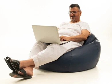 A modern and advanced technical device, expressions of focus and contemplation, accomplishing remote work tasks, a portrait of an elderly Arab Saudi Gulf man wearing pants and a t-shirt sitting on a chair between a comfortable bag using a laptop, white background.