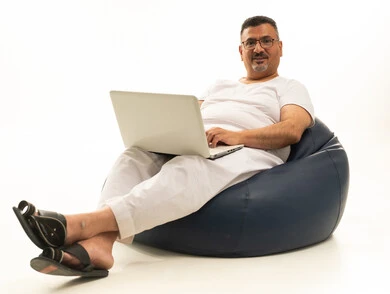 إنجاز مهام العمل عن بعد، جهاز تقني حديث ومتطور، تعابير التركيز والتمعن، بورتريه لرجل كبير بالسن عربي سعودي خليجي يرتدي سروال و فانيلة ويضع نظارات طبية يجلس على كرسي بين باج مريح يقوم باستخدام الحاسوب المحمول، خلفية بيضاء