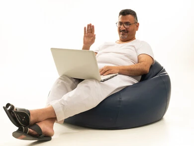 رفع اليدي بإيماءات إلقاء التحية والسلام، إجراء مكالمة فيديو، إنجاز مهام العمل عن بعد، جهاز تقني حديث ومتطور، بورتريه لرجل كبير بالسن عربي سعودي خليجي يرتدي سروال و فانيلة ويضع نظارات طبية يجلس على كرسي بين باج مريح يقوم باستخدام الحاسوب المحمول، خلفية بيضاء