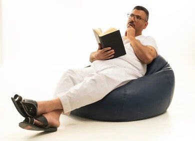 Spending time reading, learning, and culture, gestures of contemplation and focus, the concept of self-learning, a portrait of an elderly Arab Saudi Gulf man wearing pants and a t-shirt sitting on a chair between a comfortable couch reading a book, white background. Spending time reading, learning, and culture, gestures of contemplation and focus, the concept of self-learning, a portrait of an elderly Arab Saudi Gulf man wearing pants and a t-shirt sitting on a chair between a comfortable couch reading a book, white background.