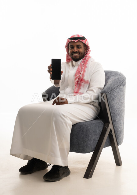 Using a mobile phone, live streaming with family and friends, browsing social media, a portrait of a Saudi Arabian Gulf man wearing traditional attire and a ghutrah holding a mobile phone in his hand, integrating modern technologies into daily life, white background.