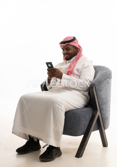 دمج التقنيات الحديثة فى الحياة اليومية ، استخدام الهاتف المحمول ، إجراء بث مباشر مع الأهل والأصدقاء، تصفح مواقع التواصل الاجتماعي ، بورتريه لرجل عربي خليجي سعودي يرتدي الثوب التقليدي والشماغ يمسك الجوال بيده ، خلفية بيضاء