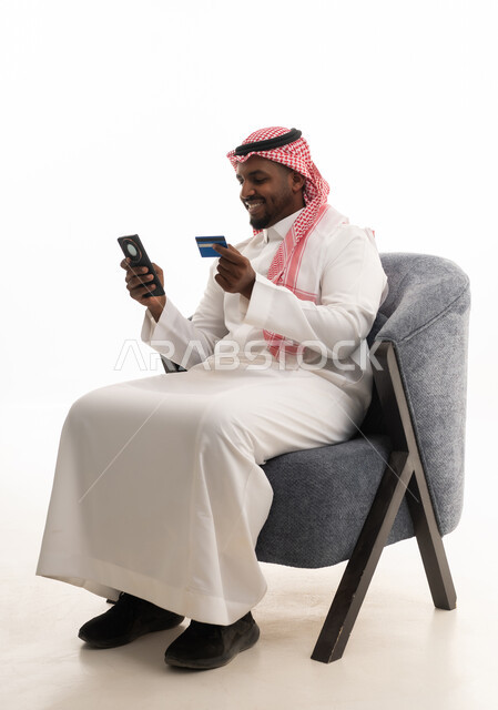 التسويق اونلاين عبر الانترنت ، الدفع عبر بطاقة الائتمان ، بورتريه لرجل عربي خليجي سعودي يرتدي الثوب التقليدي والشماغ يمسك الجوال والبطاقة بيده ،دمج التقنيات الحديثة فى الحياة اليومية ، خلفية بيضاء