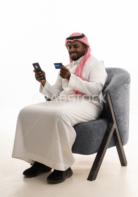 الدفع عبر بطاقة الائتمان ، التسويق اونلاين عبر الانترنت ، بورتريه لرجل عربي خليجي سعودي يرتدي الثوب التقليدي والشماغ يمسك الجوال والبطاقة بيده ،دمج التقنيات الحديثة فى الحياة اليومية ، خلفية بيضاء