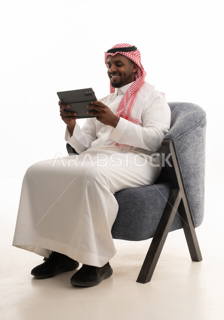 دمج التقنيات الحديثة فى الحياة اليومية ، استخدام الجهاز اللوحي  ، إجراء بث مباشر مع الأهل والأصدقاء، تصفح مواقع التواصل الاجتماعي ، بورتريه لرجل عربي خليجي سعودي يرتدي الثوب التقليدي والشماغ يمسك جهاز التابلت بيده ، خلفية بيضاء