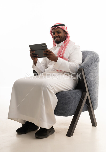 تصفح مواقع التواصل الاجتماعي ، بورتريه لرجل عربي خليجي سعودي يرتدي الثوب التقليدي والشماغ يمسك جهاز التابلت بيده ، دمج التقنيات الحديثة فى الحياة اليومية ، استخدام الجهاز اللوحي  ، إجراء بث مباشر مع الأهل والأصدقاء،خلفية بيضاء