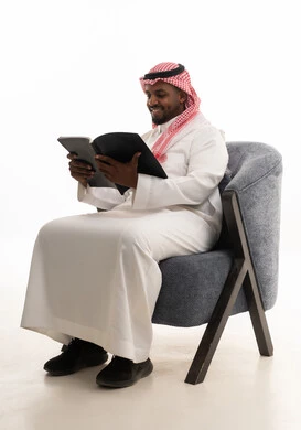 بورتريه لرجل عربي خليجي سعودي يرتدي الثوب التقليدي والشماغ يجلس على مكتب يقوم بقراءه كتاب   ، حب المطالعة والقراءة ،تمضية أوقات الفراغ بالتعلم والتثقف الذاتي, إيماءات التأمل والتفكير،خلفية بيضاء