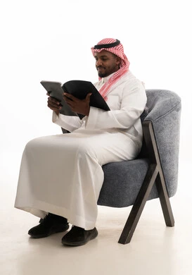 إيماءات التأمل والتفكير، بورتريه لرجل عربي خليجي سعودي يرتدي الثوب التقليدي والشماغ يجلس على مكتب يقوم بقراءه كتاب   ، حب المطالعة والقراءة ،تمضية أوقات الفراغ بالتعلم والتثقف الذاتي,خلفية بيضاء