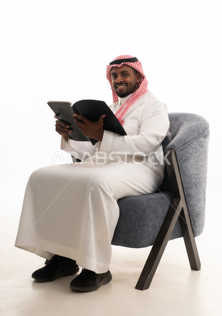 تمضية أوقات الفراغ بالتعلم والتثقف الذاتي, حب المطالعة والقراءة ،إيماءات التأمل والتفكير، بورتريه لرجل عربي خليجي سعودي يرتدي الثوب التقليدي والشماغ يجلس على مكتب يقوم بقراءه كتاب   ، خلفية بيضاء