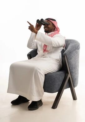 The love of exploration and adventure, using binoculars to discover distant things and bring them closer, a portrait of an Arab Gulf Saudi man wearing traditional attire and a ghutrah sitting on a comfortable chair holding binoculars in his hand and looking at something, gestures of enjoyment and pleasure, white background.