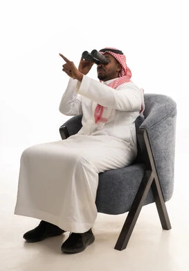 إيماءات الاستمتاع والسرور، استعمال المنظار للكشف عن الاشياء البعيدة وتقريبها, بورتريه لرجل عربي خليجي سعودي يرتدي الزي التقليدي والشماغ يجلس على كرسي مريح يحمل بيده المنظار ثنائي العينين وينظر الى شيء ما، حب الاستكشاف والمغامرة، خلفية بيضاء