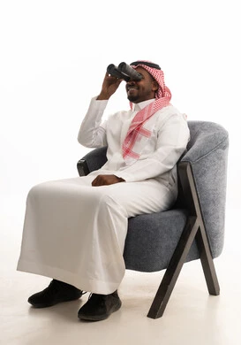 A portrait of a Saudi Arabian Gulf man wearing traditional attire and a ghutrah sitting on a comfortable chair holding a binocular in his hand and looking at something, a love for exploration and adventure, gestures of enjoyment and pleasure, using the binoculars to reveal distant objects and bring them closer, with a white background.