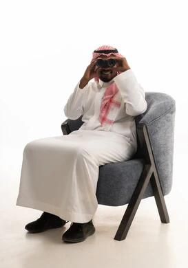 استعمال المنظار للكشف عن الاشياء البعيدة وتقريبها, بورتريه لرجل عربي خليجي سعودي يرتدي الزي التقليدي والشماغ يجلس على كرسي مريح يحمل بيده المنظار ثنائي العينين وينظر الى شيء ما، حب الاستكشاف والمغامرة، إيماءات الاستمتاع والسرور، خلفية بيضاء