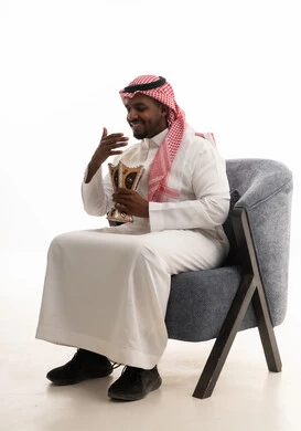 الاهتمام بالمظهر الخارجي والأناقة, التعطر والتطيب بالبخور, استخدام خشب العود بالأعياد والمناسبات, العادات والتقاليد السعودية, بورتريه لرجل عربي خليجي سعودي يرتدي الثوب التقليدي والشماغ  يجلس على كرسي مريح يقوم بالتعطر من المبخرة,خلفية بيضاء