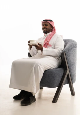 بورتريه لرجل عربي خليجي سعودي يرتدي الثوب التقليدي والشماغ  يجلس على كرسي مريح يقوم بشرب القهوة، إيماءات السعادة والسرور، قضاء وقت الفراغ باحتساء المشروب المفضل، تناول الأعشاب الشتوية الدافئة، الاستمتاع بالمشروبات الساخنة،خلفية بيضاء