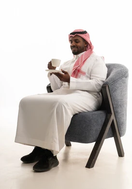 Enjoying warm winter herbs, savoring hot beverages, spending leisure time sipping a favorite drink, a portrait of a Saudi Arabian Gulf man wearing traditional attire and a ghutrah sitting on a comfortable chair drinking coffee, gestures of happiness and joy, white background.