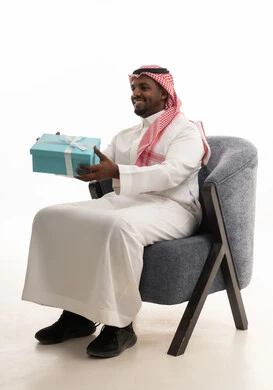 بورتريه لرجل عربي خليجي سعودي يرتدي الثوب التقليدي والشماغ  يجلس على كرسي مريح يحمل بيديه صندوق هدية, ايماءات السرور والسعادة، مفهوم الشكر والامتنان، تقديم الهدايا بالاعياد والمناسبات الخاصة، التعبير عن الحب والتقدير والاحترام،خلفية بيضاء