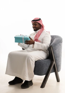 التعبير عن الحب والتقدير والاحترام،بورتريه لرجل عربي خليجي سعودي يرتدي الثوب التقليدي والشماغ  يجلس على كرسي مريح يحمل بيديه صندوق هدية, ايماءات السرور والسعادة، مفهوم الشكر والامتنان، تقديم الهدايا بالاعياد والمناسبات الخاصة، خلفية بيضاء